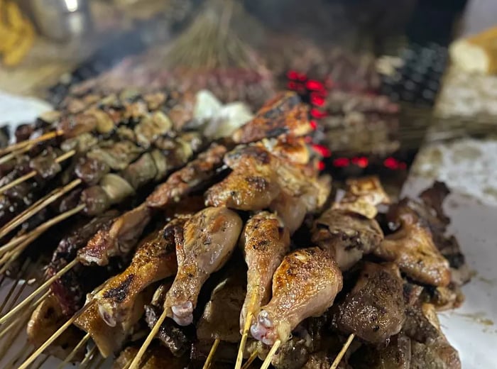 A pile of grilled meat skewers placed beside a grill with more meat cooking.