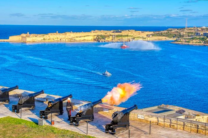 At noon, a cannon is fired from the Saluting Battery in Valletta, Malta, with Birgu visible in the background.