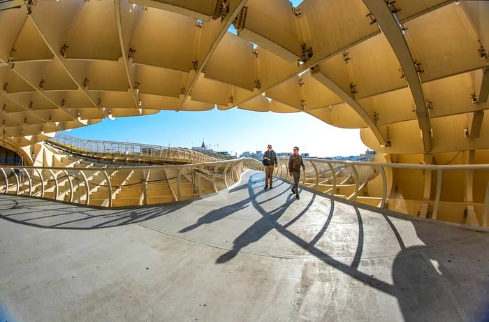 Two people walk along an elevated pathway beneath a wooden lattice structure overhead.