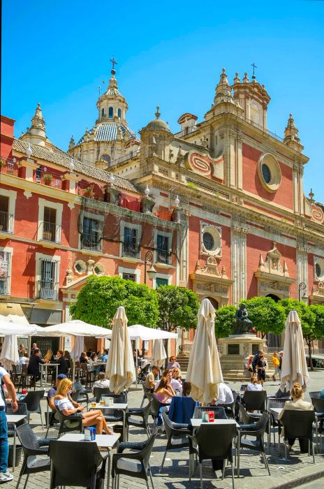 Rows of tables shaded by umbrellas in front of a stunning baroque church, featuring a pale-pink facade and numerous turrets