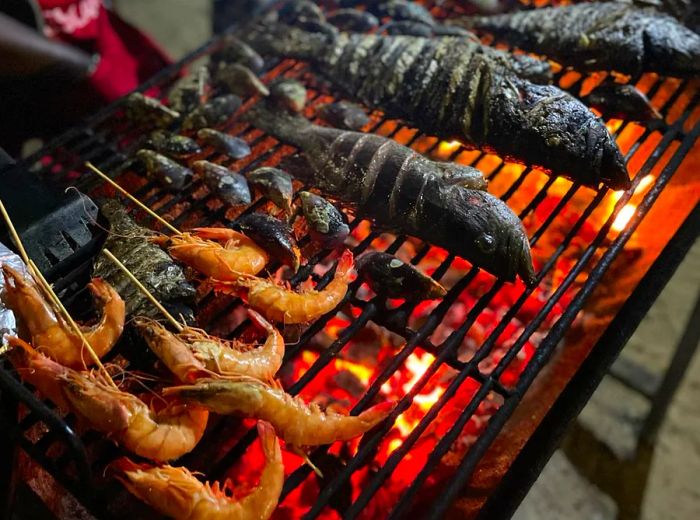 Shrimp skewers and whole, charred fish sizzling on a vibrant coal grill.