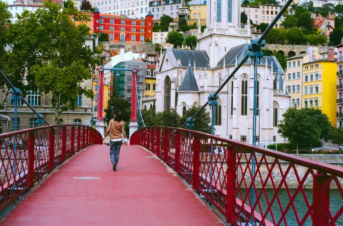 A woman strolls across a red footbridge spanning the Saône River in Lyon