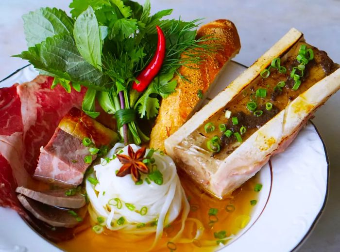 A bowl of pho featuring a swirl of noodles, dark slices of meat, a bone filled with marrow, a bundle of herbs, and chili peppers.