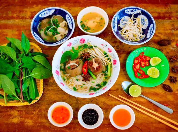 A hearty bowl of pho brimming with meats, fresh herbs, and noodles, accompanied by an array of toppings and sauces placed around the bowl.