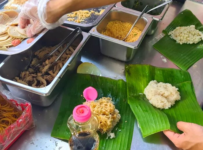 A chef carefully assembles a packet of sticky rice, selecting ingredients from the prep trays and wrapping them in a banana leaf.