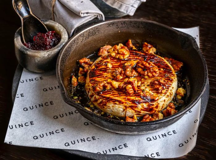 A round of cheese elegantly presented in a pan, drizzled with dark sauce, sprinkled with nuts, and served alongside a basket of bread.