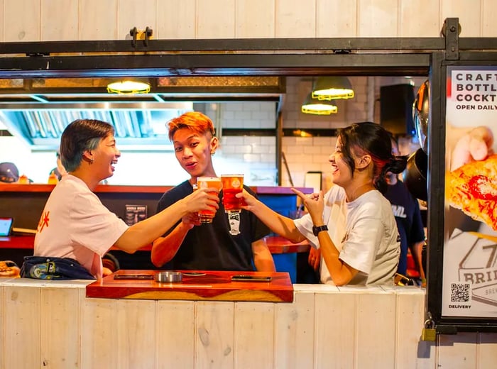 Three patrons toast with beers through an open window, while a pizza advertisement is visible outside.