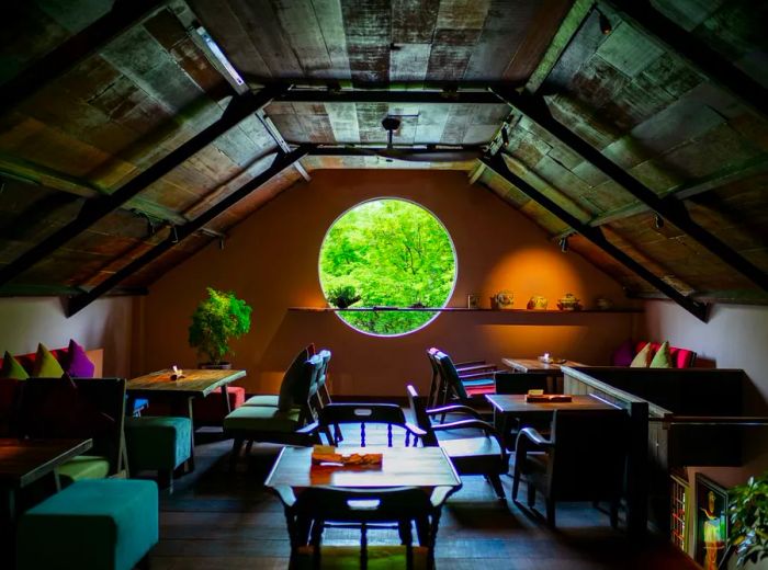 A dimly lit dining space, illuminated by a large round window, featuring low seating and benches with colorful cushions surrounding wooden tables.