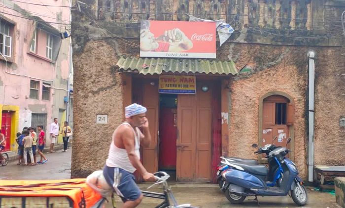 The exterior of a restaurant, viewed through the hustle of passersby, featuring a Coca-Cola advertisement above its entrance.