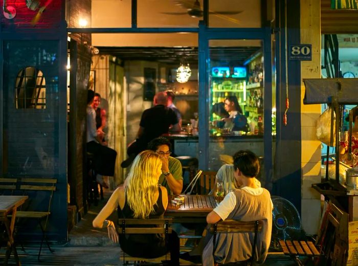 Patrons enjoy drinks at outdoor tables beneath a branded awning in front of the open bar.