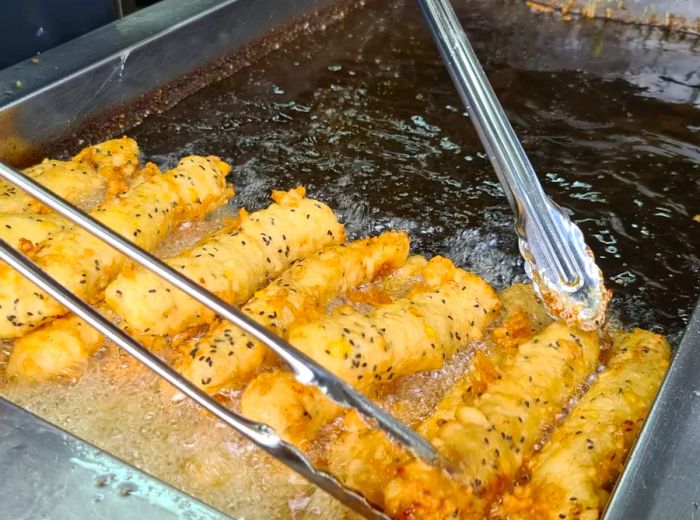 Bananas being battered and fried in a large pot of oil, with tongs poised above.