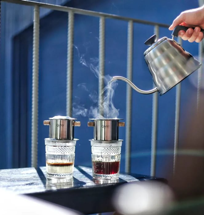 A hand pours hot water from a pot into a phin-style coffee brewer placed on a windowsill, next to another brewing device.