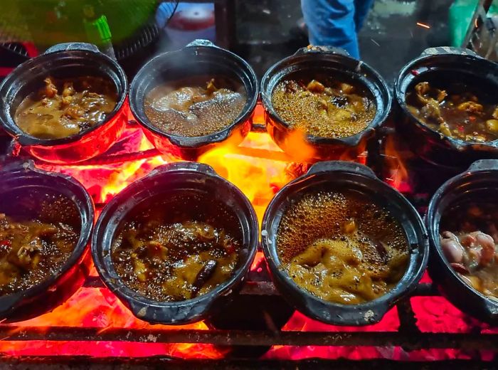 Small iron pots simmering with dark broth placed on a grate over a coal fire.