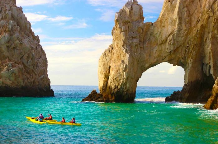 Individuals enjoying a kayak ride in vibrant yellow kayaks, equipped with life vests, glide along the stunning stone arc landmark in the beautiful turquoise waters of Cabo San Lucas beneath a clear blue sky.