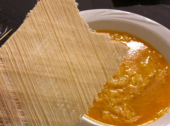 A flat sheet of pulled noodles rests on the edge of a bowl filled with orange-tinted broth.