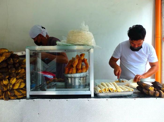 Two chefs busily prepare banana lumpia behind a counter, with freshly made pieces displayed for customers.