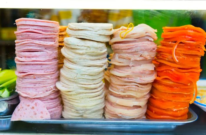 Four stacks of cold cuts displaying various colors and textures arranged on a metal tray.