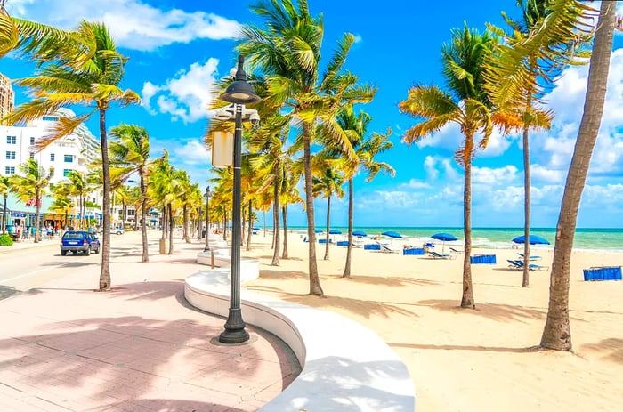 The beachfront promenade in Fort Lauderdale is adorned with palm trees, while blue sun loungers dot the pristine white-sand beach just beyond.