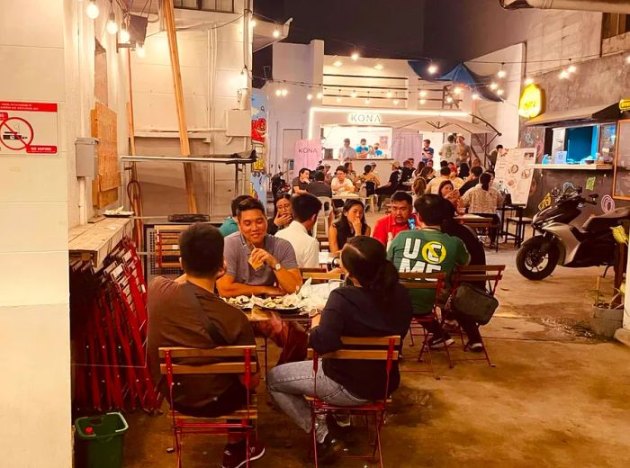 Patrons enjoy their beers at small picnic tables in a concrete area adorned with vibrant neon signs.