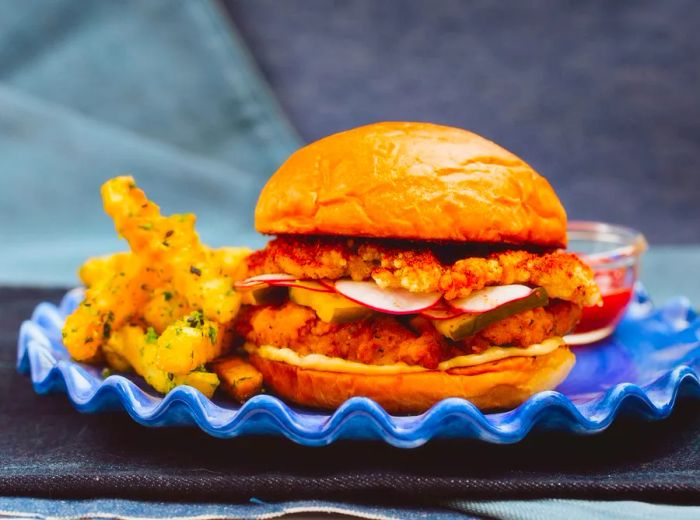 A sandwich featuring two hefty fried chicken pieces, crisp radishes, and a flavorful sauce, accompanied by a serving of herb-seasoned fries.