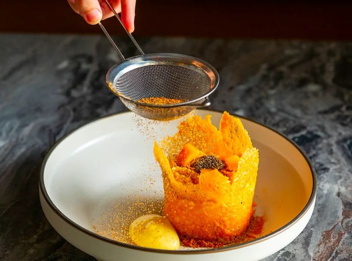 A chef delicately sprinkles a topping through a fine mesh strainer onto an elaborately presented dessert housed in a crackly edible shell.