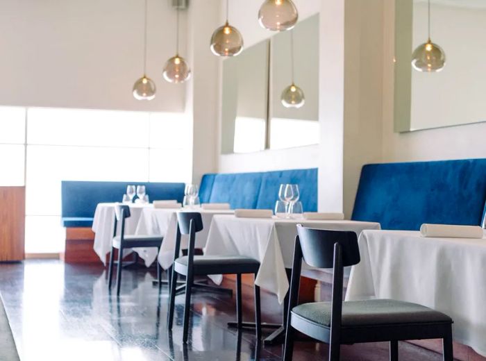 The interior of the café is bright, featuring a light blue banquette, a long mirror adorning the wall, crisp white tablecloths, and stylish metallic pendant lights.