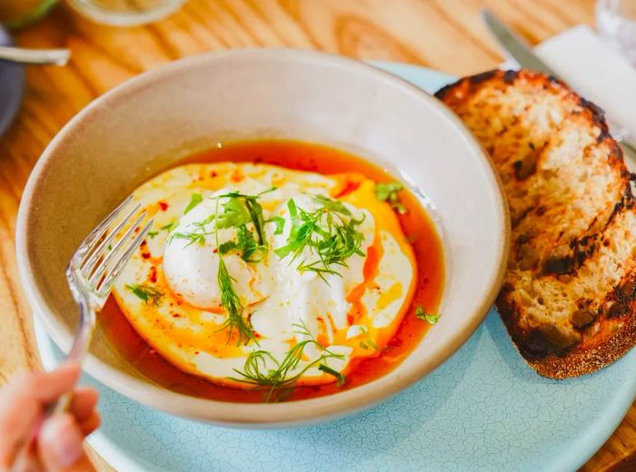 A bowl of soft-boiled eggs in a vibrant red sauce, accompanied by slices of toast.