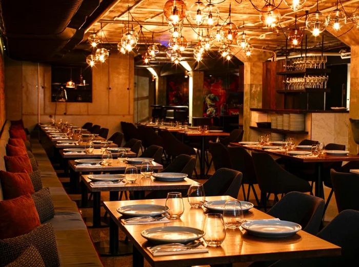 A dimly lit dining area featuring a collection of exposed bulbs hanging from the ceiling, with tables aligned against a lengthy banquette.