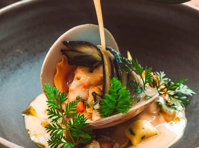 A chef drizzles sauce over a large clam adorned with herbs and garnishes.