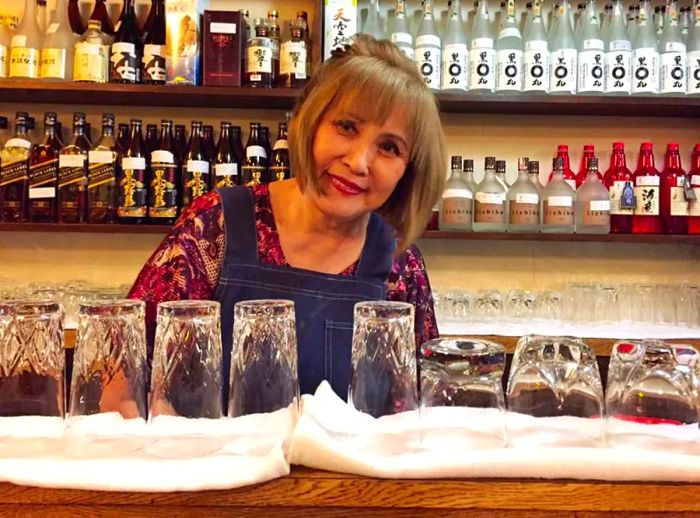 A woman stands behind a bar adorned with drying drinkware, surrounded by bottles from Japan.