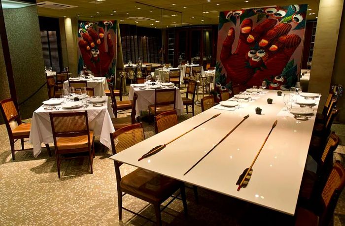 The dining room features large photographs of hands holding small dishes, elegantly set white tablecloths, and a prominent communal table decorated with long arrows in the foreground.