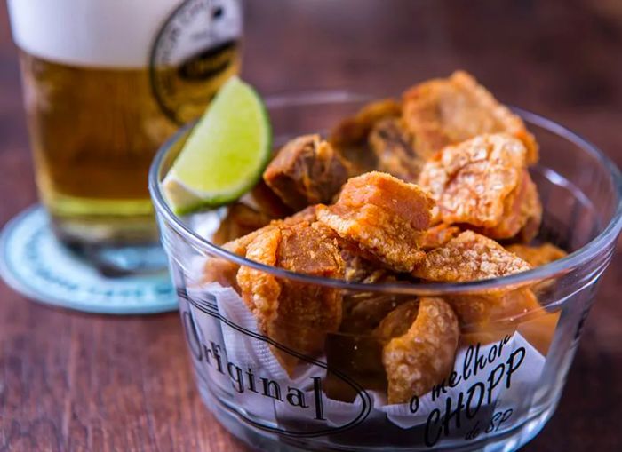 A glass bowl labeled Original, filled with chicharron and a lime wedge, with a blurred glass of beer in the background.