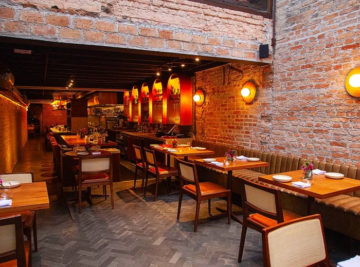 The restaurant boasts an inviting atmosphere with exposed brick walls, an array of warm lighting fixtures, and light wooden tables complemented by a soft brown leather banquette.