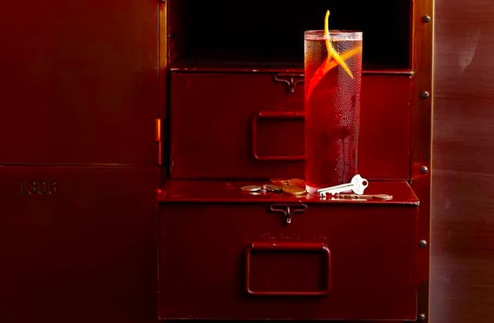 A highball glass sits on the open door of a secure box, with a key and some coins resting nearby.