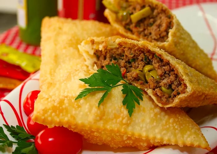 A paper plate holds two halves of a meat-filled pastry, one cut open to reveal its filling, alongside an intact deep-fried pastry, garnished with herbs and cherry tomatoes.