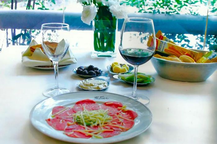 Thinly sliced meat is beautifully arranged on a plate, accompanied by two glasses of wine, an assortment of small plates, bread baskets, and a vase of flowers, all set on a sunny outdoor patio.