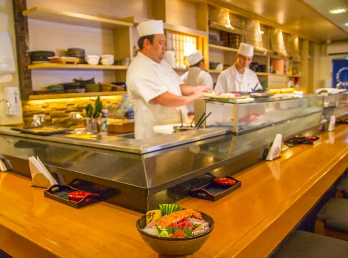 A vibrant bowl of chirashi brimming with assorted fish pieces rests on an L-shaped sushi counter, where chefs skillfully prepare dishes behind the bar.