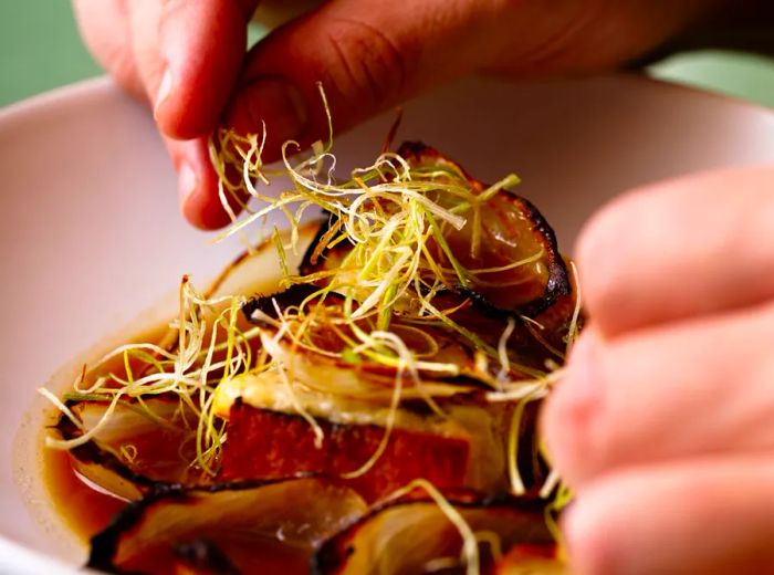 A chef's hands prepare a dish featuring crispy chive roots atop a soup filled with onions, bread, and melted cheese, with a hint of broth at the base.