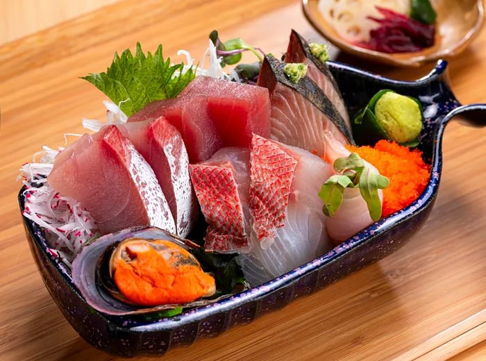 A small dish brims with an assortment of fish slices, a mussel, a cluster of roe, and a mound of wasabi, all presented on a wooden tray alongside other slightly blurred small plates.