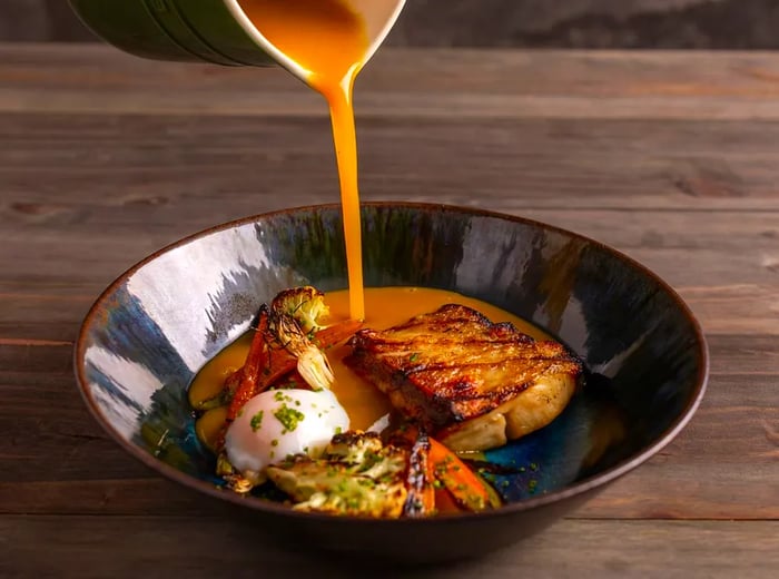 An unseen server gracefully pours broth from a pitcher into a bowl containing tambaqui fish fillet, cauliflower, carrots, and a poached egg. The glazed bowl rests on a dark wooden table.