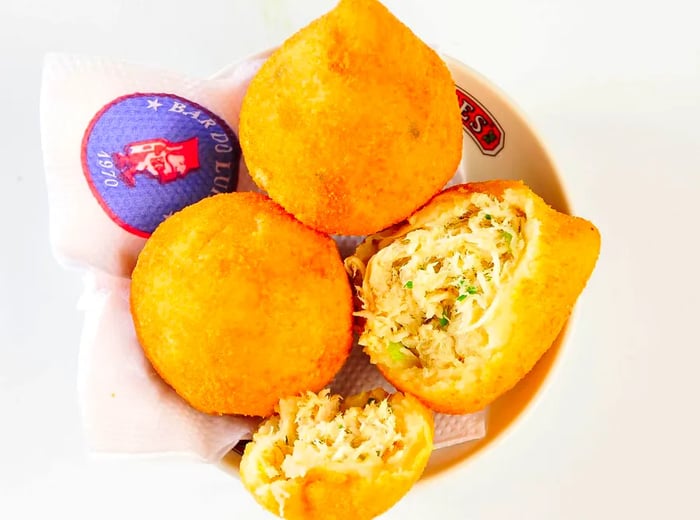 A plate of chicken fritters, featuring two whole pieces and one sliced open, rests on a napkin from Bar do Luiz Fernandes against a plain background.