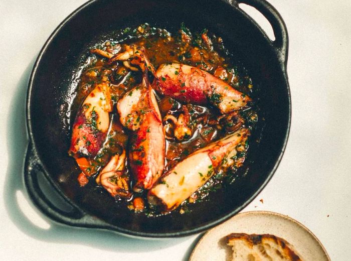 A cast iron skillet holds squid in a savory sauce, with a side of bread.