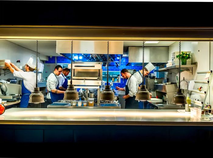 A kitchen team operates in an open space filled with steel appliances and a bright backsplash, all contrasting with the darker dining area, viewed through a cutout.