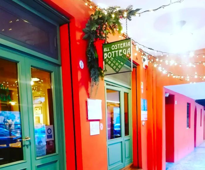 The exterior of a restaurant under a portico, adorned with a hanging sign, twinkling lights, and lush greenery at the entrance.