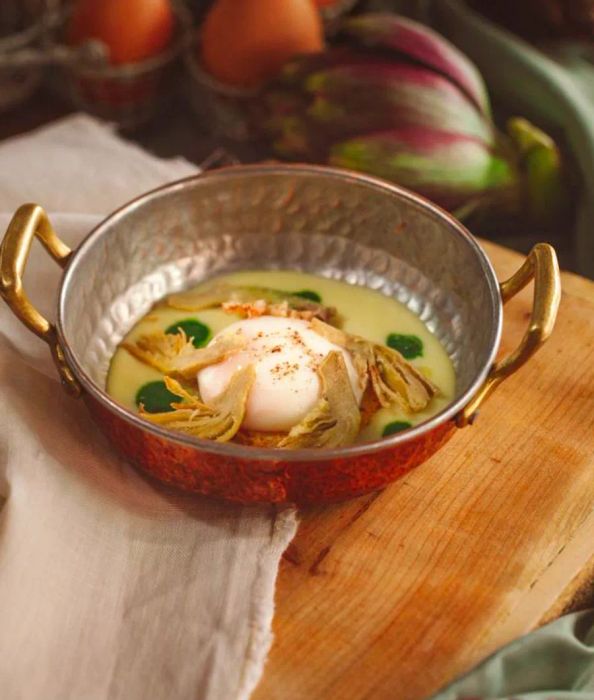 A soft-boiled egg nestled in a creamy potato blend within a hammered silver pot, elegantly paired with artichokes.