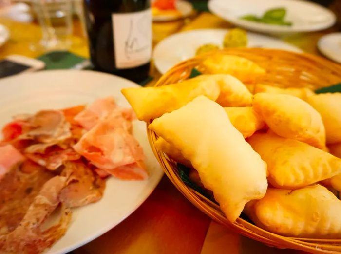 Crescentine fritte presented in a wooden basket next to a platter of sliced meats.