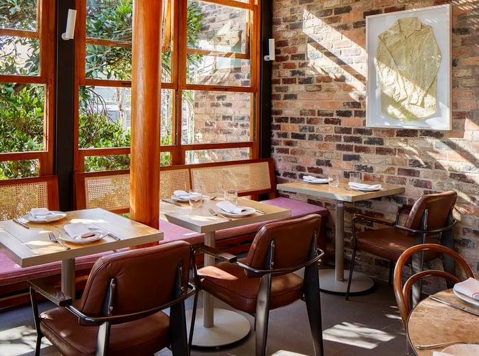 A bright, spacious dining area featuring cane banquettes and modern leather chairs, with tables set for lunch and greenery visible outside.