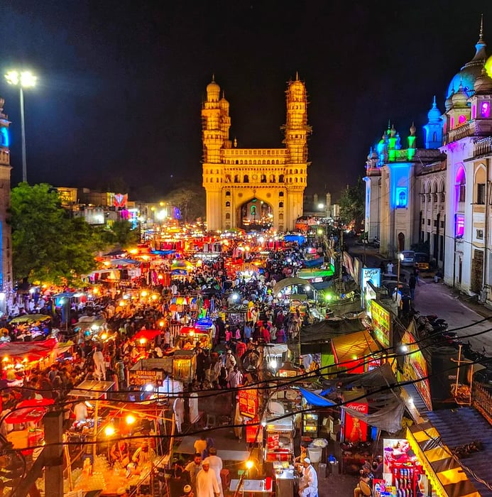 A panoramic view of nighttime street stalls with the illuminated Charminar in the background.