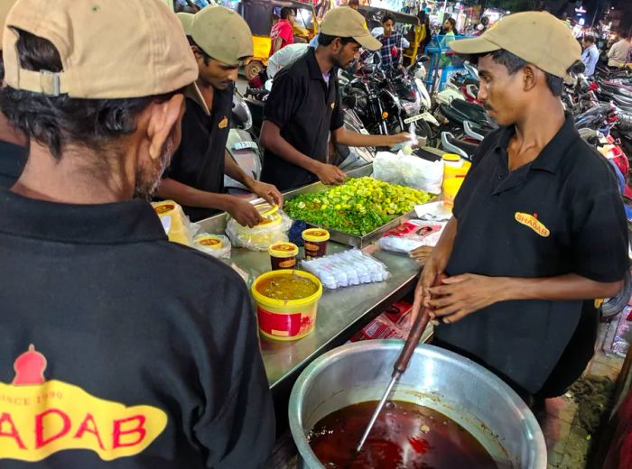 Workers prepare bowls at a table laden with toppings in a bustling night market.