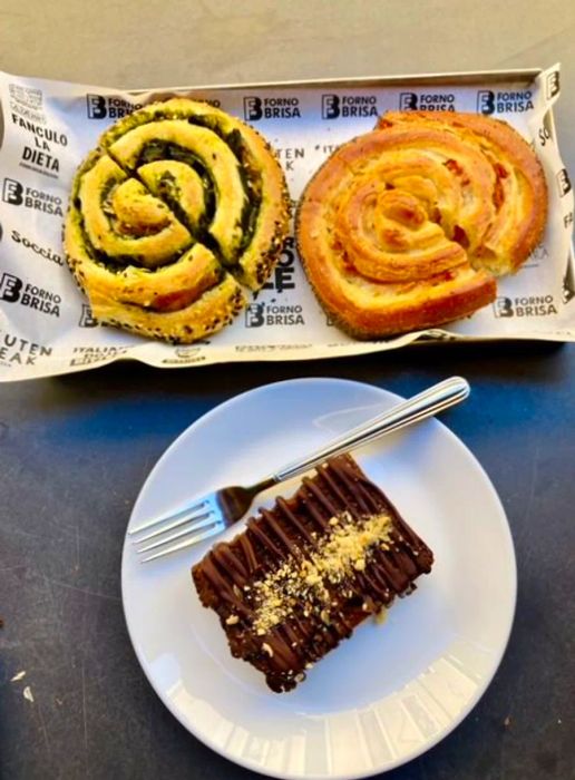 A tray lined with paper showcases two swirled pastries—one filled with green and the other with pale orange—alongside a plate featuring a slice of chocolate cake adorned with crumbled nuts.